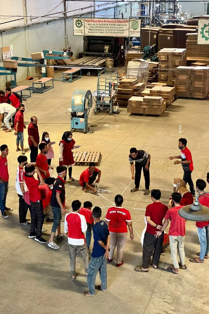 The whole JMP crew forming a wide circle on the production floor, among stacks of carton blanks and machinery, watching a game unfold.