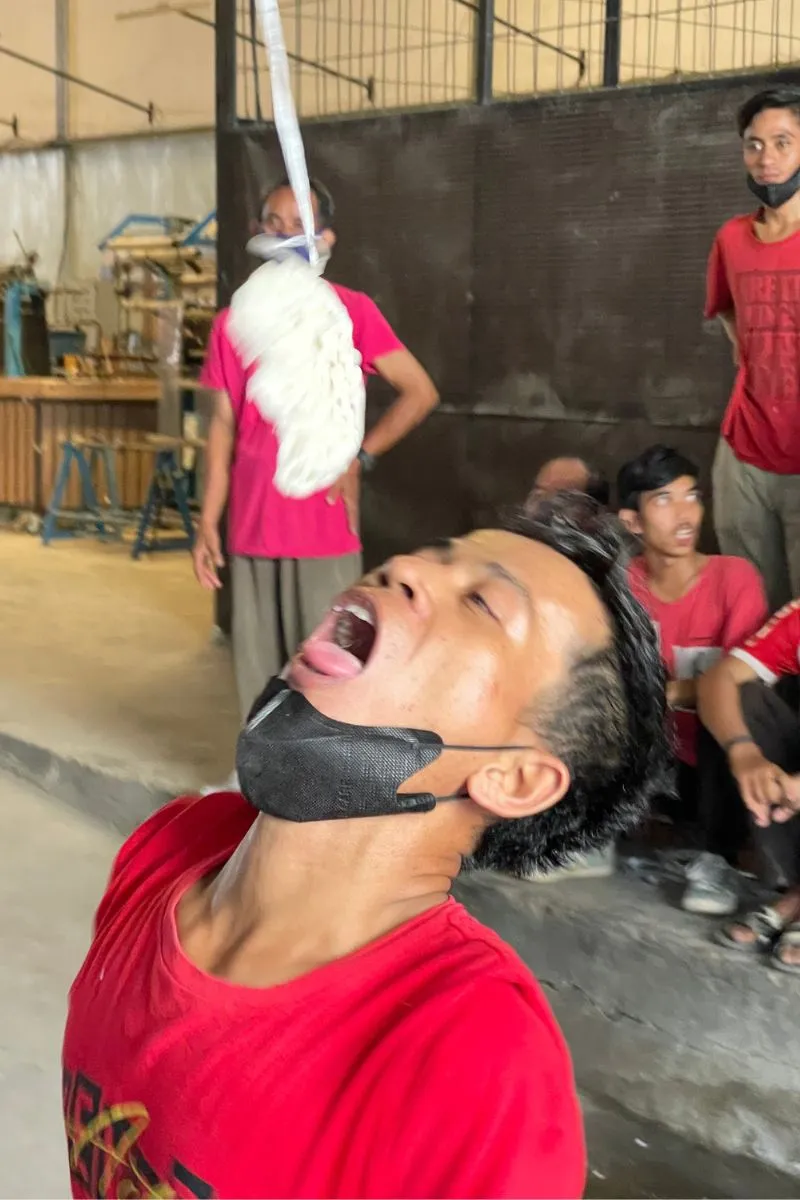 The kerupuk-eating contest on the production floor — a contestant tilting his head back with his mask pulled under his chin.