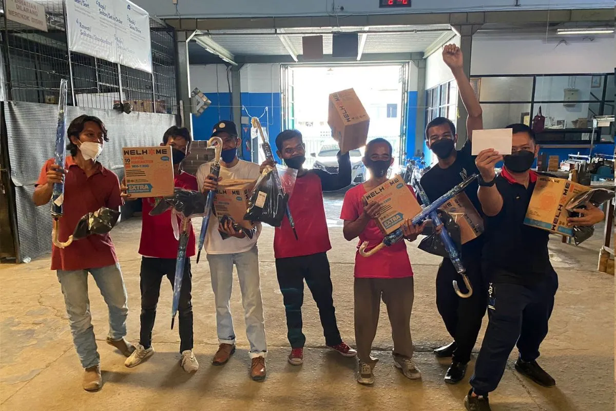 Winners carrying home umbrellas and boxed household appliances, posing at the factory door.