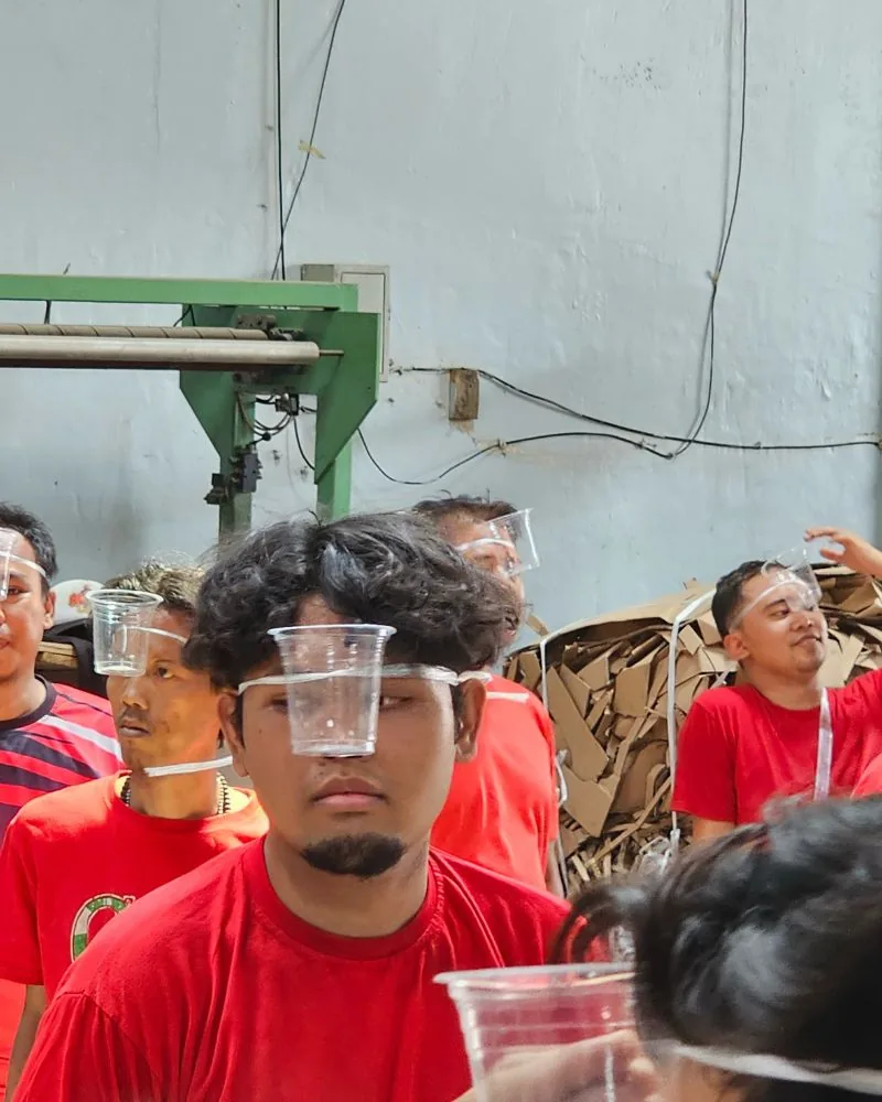 Peserta lomba 17-an JMP dengan gelas plastik di wajah, suasana meriah di antara mesin produksi.