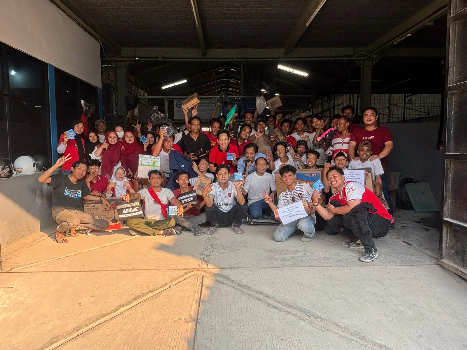 Foto bersama tim JMP diambil dari luar, terlihat melalui bukaan pintu gudang dengan lantai beton yang kena sinar matahari.