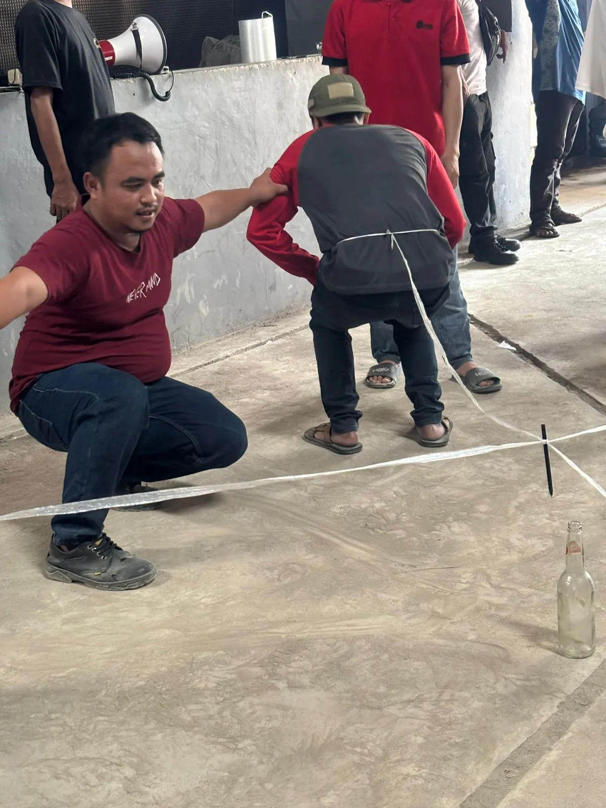 Lomba memasukkan paku ke dalam botol — paku diikat tali di pinggang peserta, botol kaca berdiri di lantai beton.