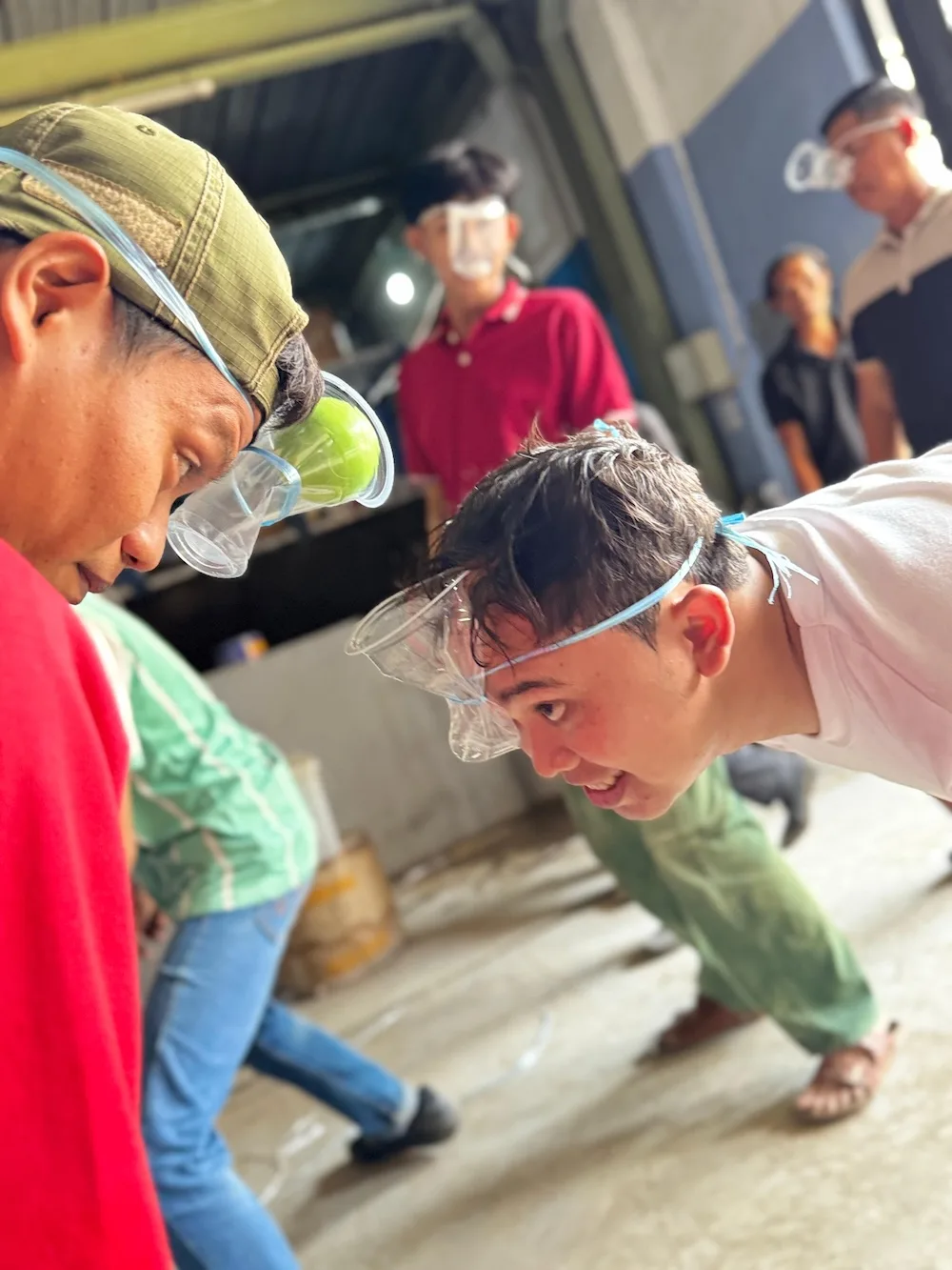 Dua peserta dengan cangkir plastik terikat di muka, satu cangkir sudah berisi bola hijau, di dekat pintu gudang yang terbuka.