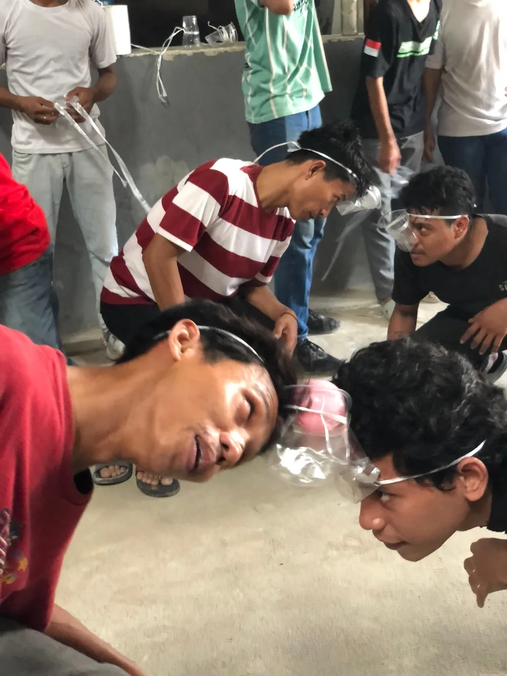 Peserta berlutut di lantai dengan cangkir plastik terikat di kepala, memindahkan balon tanpa menyentuhnya dengan tangan.