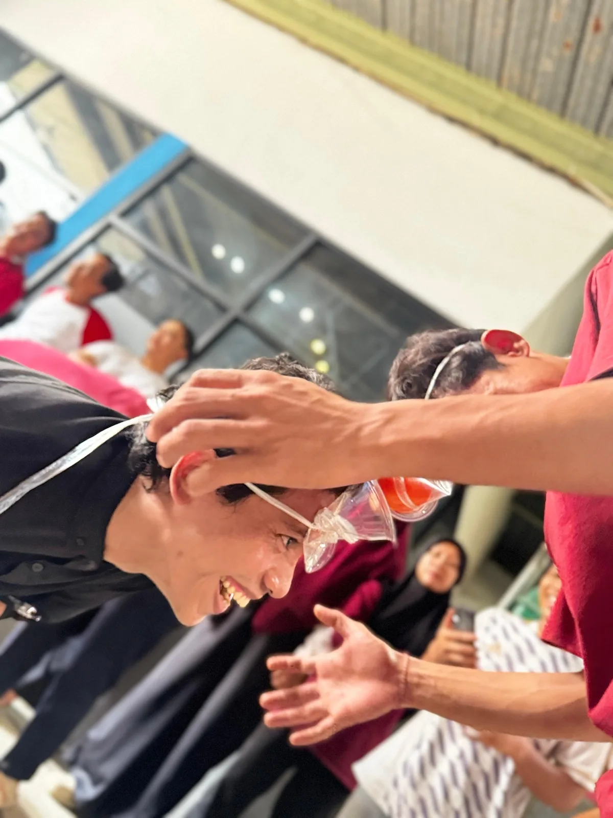Peserta tertawa saat memindahkan air berwarna antar cangkir plastik yang diikat ke muka, ditonton rekan-rekannya.