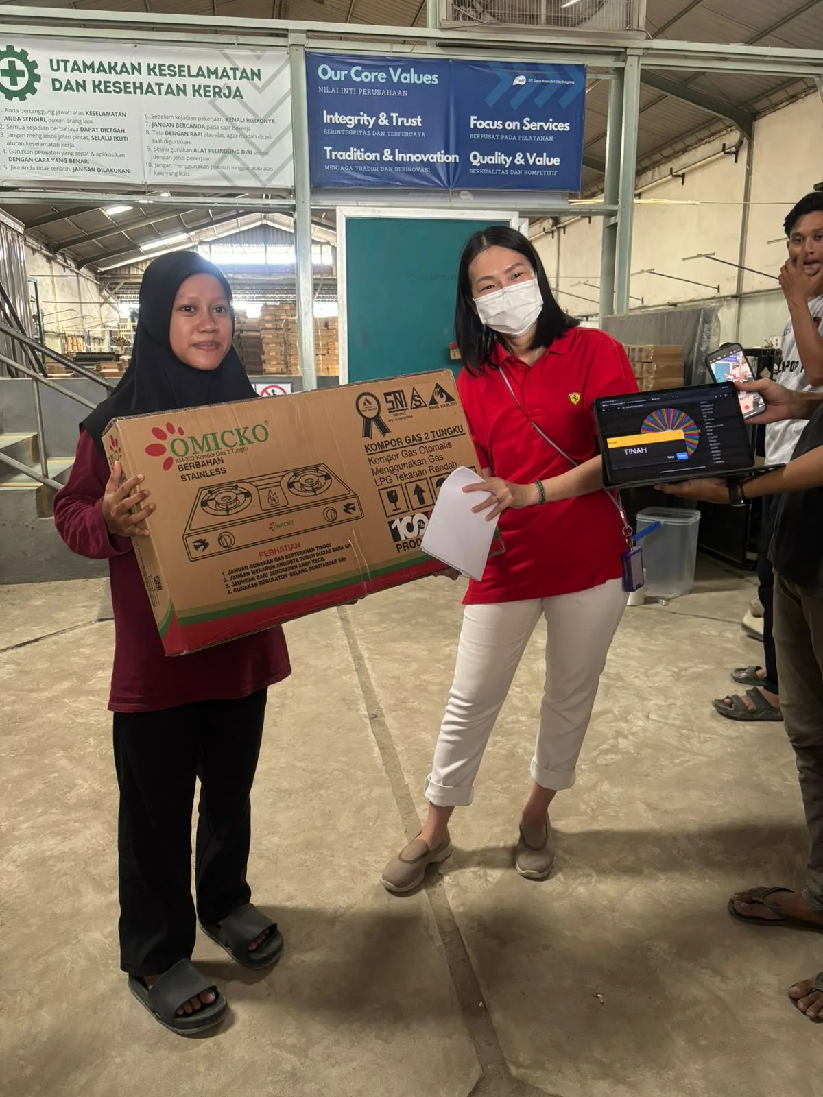 Pembagian hadiah: kompor gas dua tungku diangkat di depan banner Nilai Inti Perusahaan, di sebelahnya laptop menampilkan roda undian.