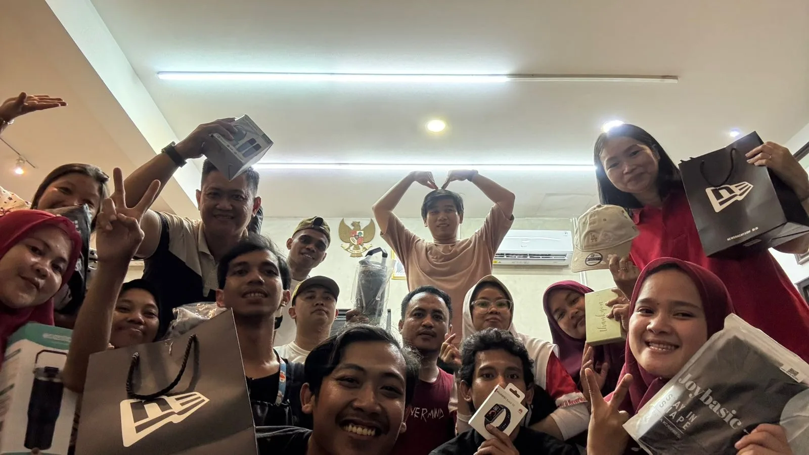 Wefie tim JMP di ruang kantor sambil memegang hadiah — tas, topi, dan tumbler — di bawah lambang Garuda Pancasila.