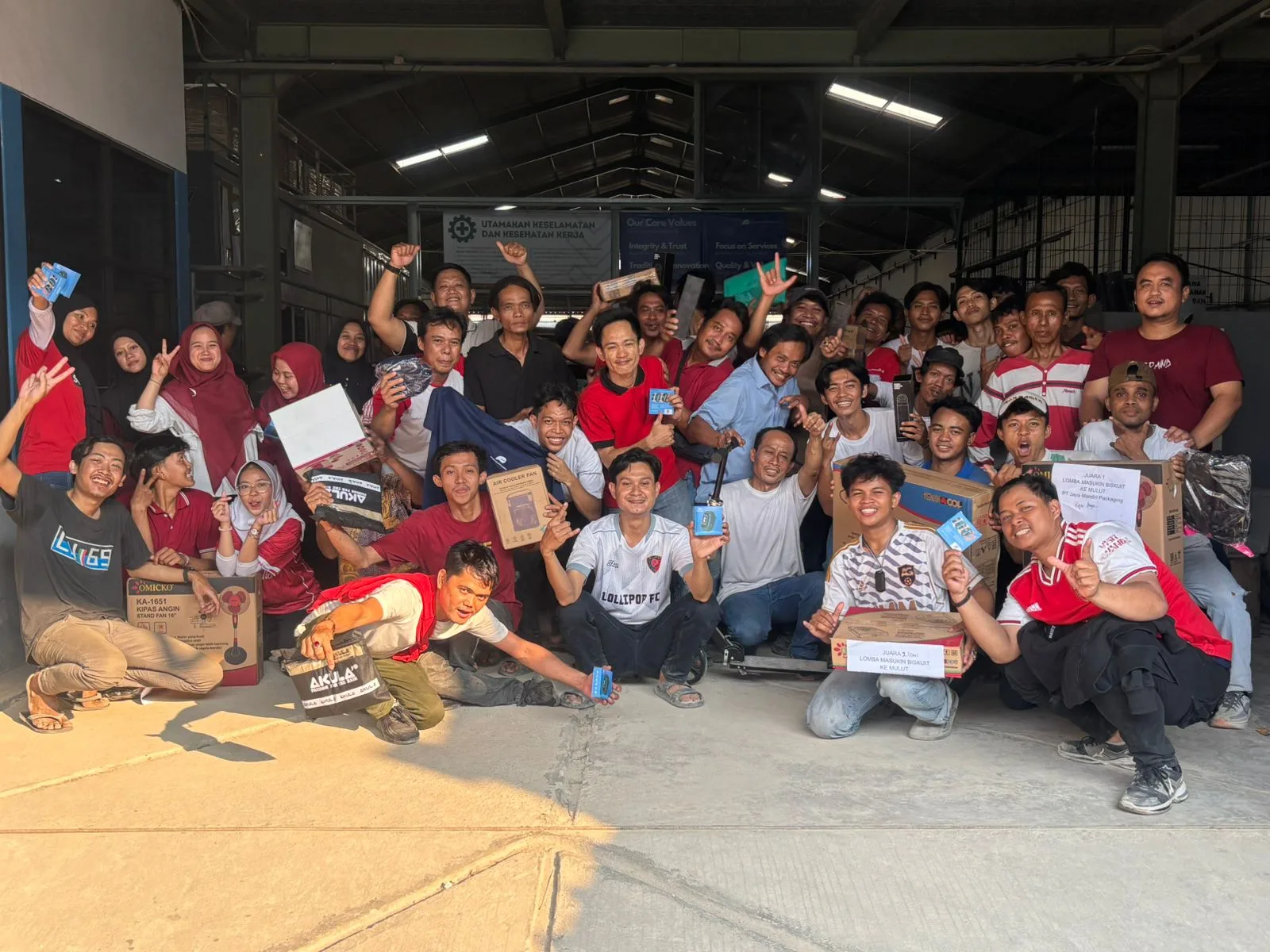 Foto bersama seluruh tim JMP memegang hadiah lomba HUT RI ke-80, dengan papan 'Juara 1 Lomba Masukin Biskuit ke Mulut' di barisan depan.