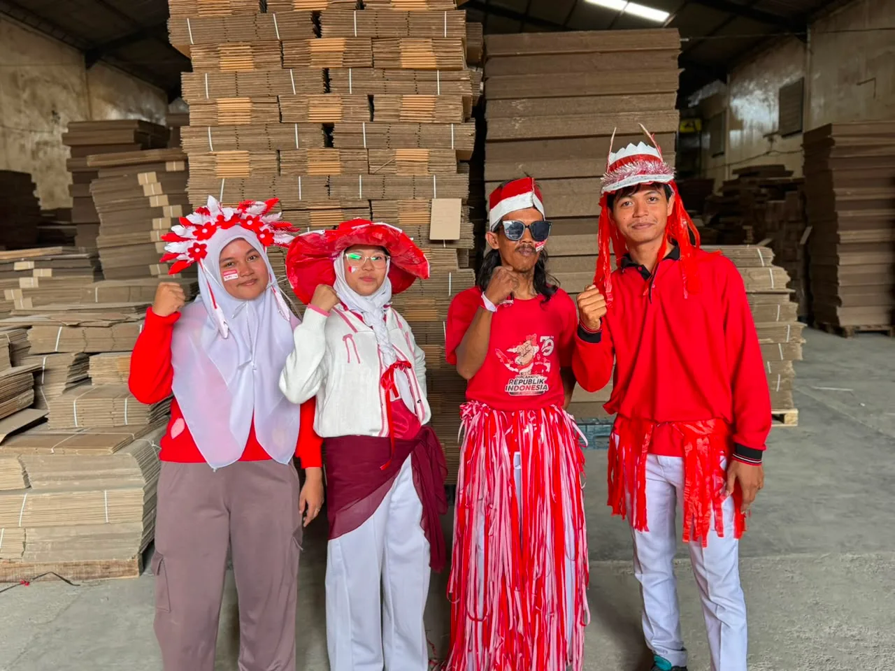 Empat karyawan berkostum merah putih berjejer di depan tumpukan lembaran karton — topi lebar dari plastik merah, rok dari untaian plastik merah putih.