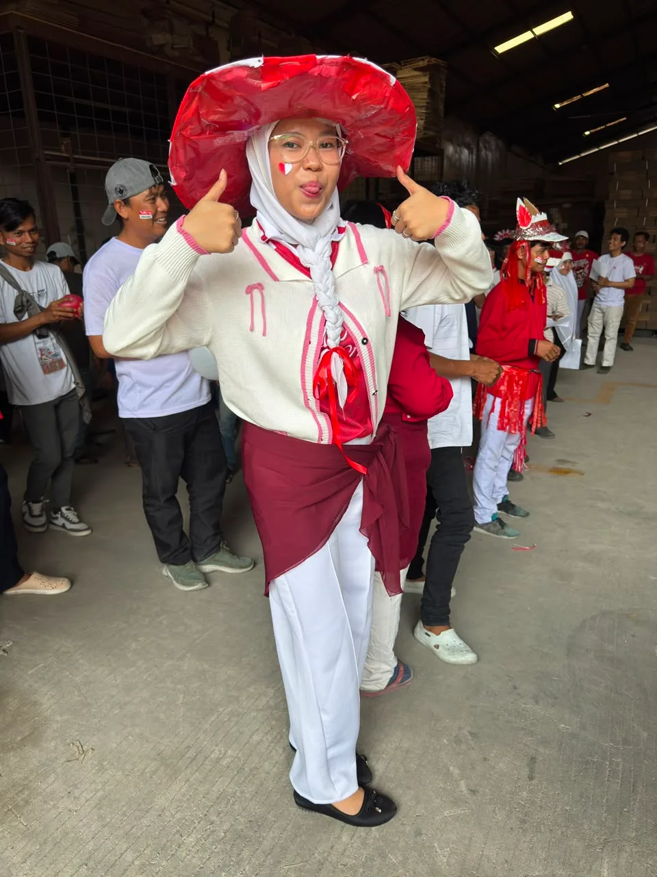 Juara 3 dress code berpose dengan dua jempol, memakai topi lebar dari plastik merah dan selempang merah putih, di lorong gudang yang penuh peserta lain.