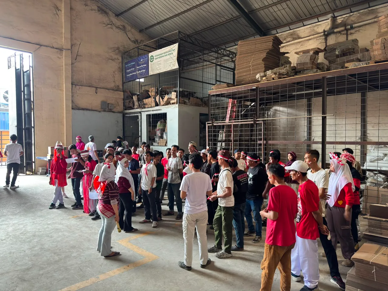 Peserta berkumpul untuk pembagian tim di lantai gudang, di bawah papan Sasaran Keselamatan dan Kesehatan Kerja dan rak berisi lembaran karton.