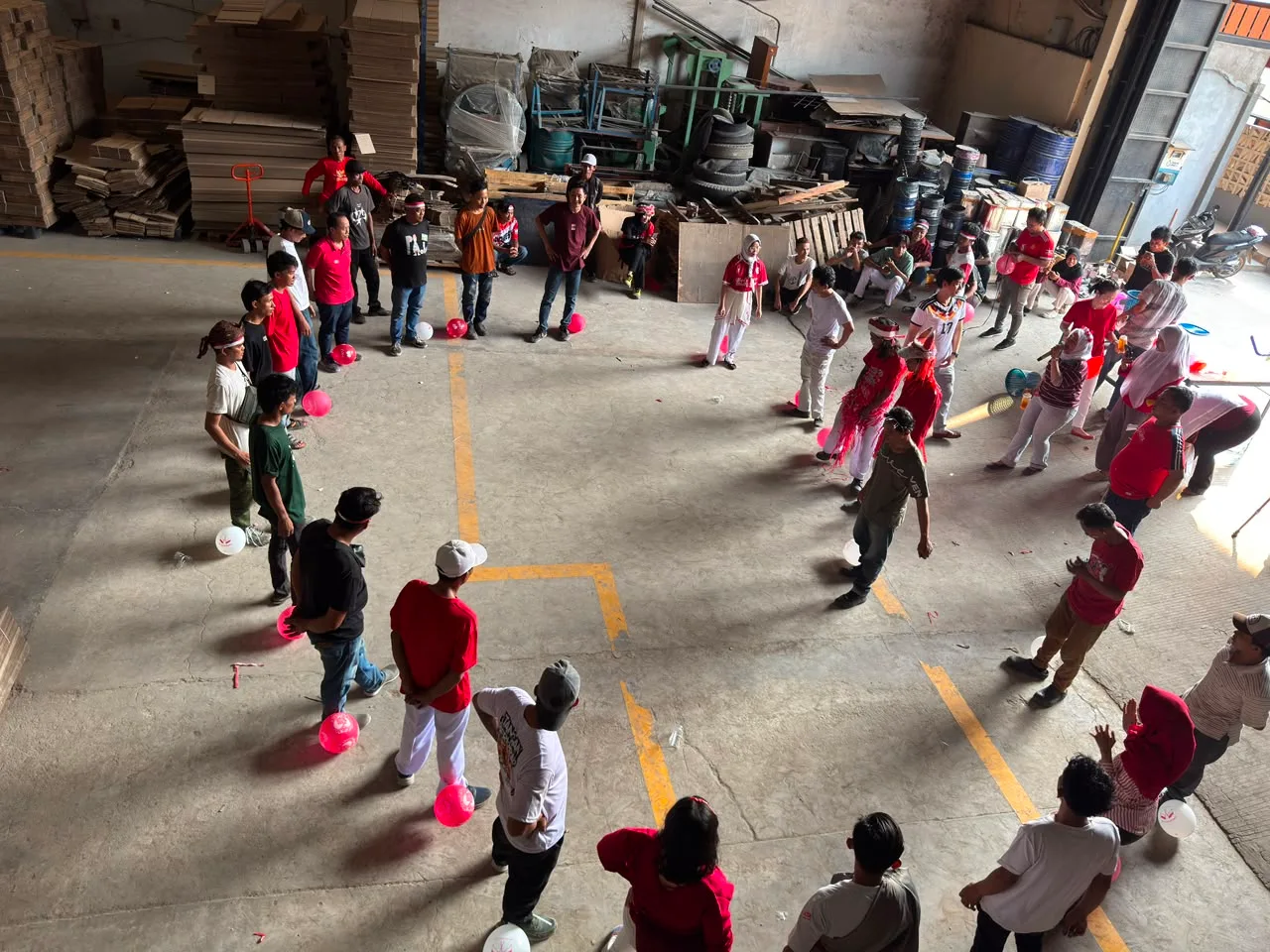 Peserta berdiri melingkar lebar di lantai gudang, masing-masing dengan balon merah muda di dekat kaki, bersiap untuk lomba injak balon.
