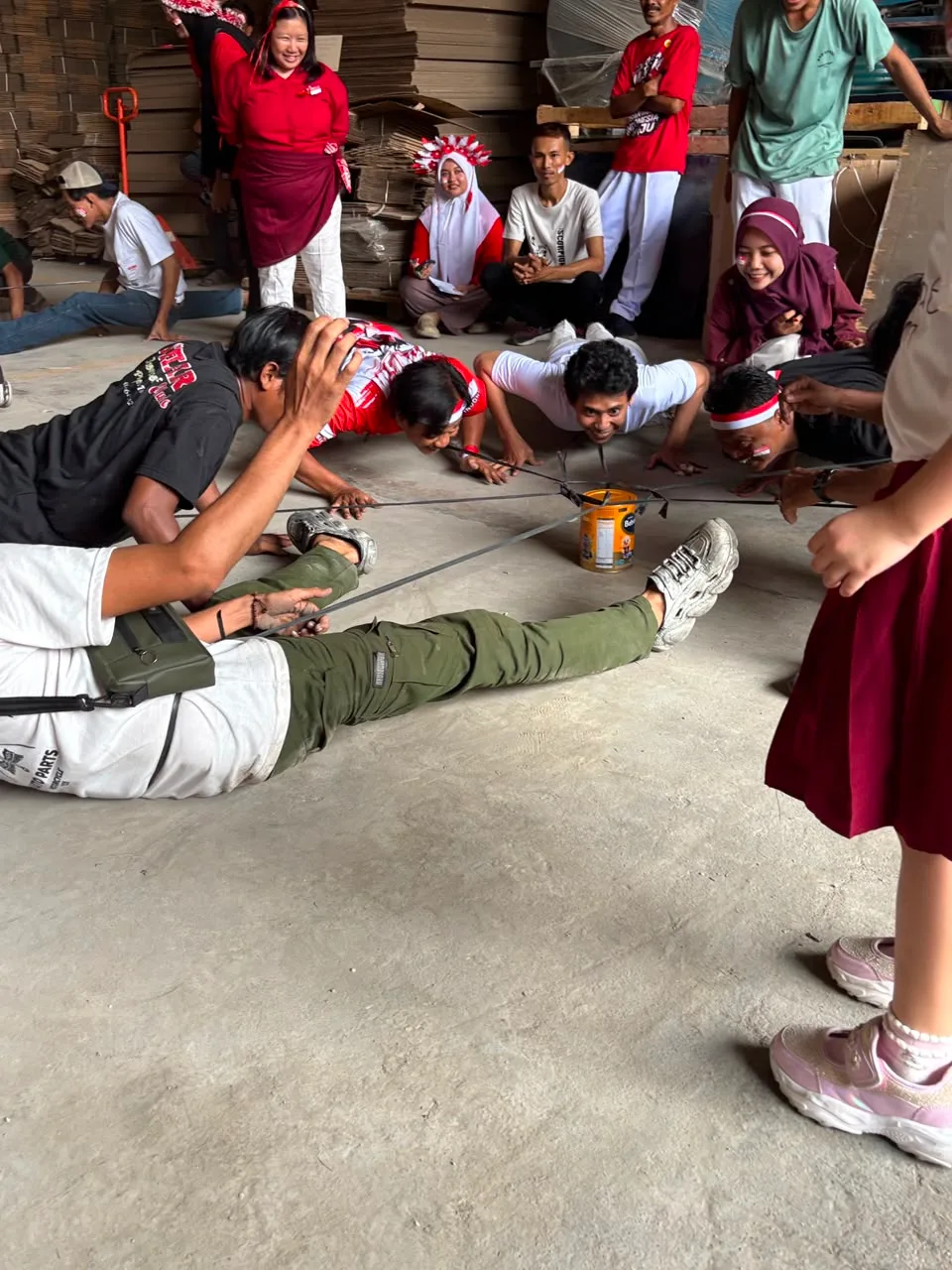 Lima peserta tiarap di lantai beton, masing-masing menarik satu tali yang diikat ke kaleng di tengah, ditonton rekan-rekan yang duduk di belakang.