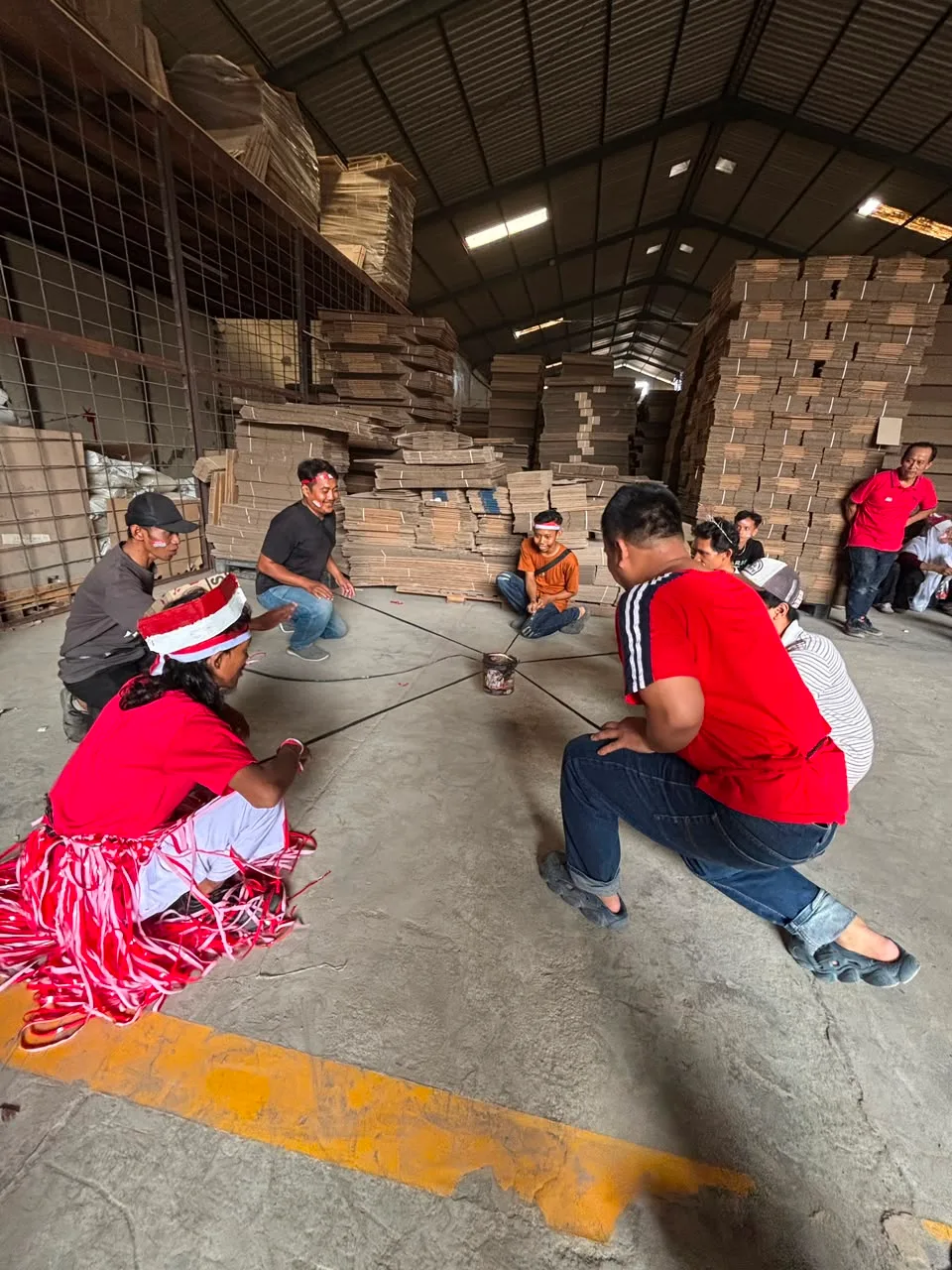 Regu lain mengangkat kaleng yang sama sambil berjongkok, lima tali merentang dari kaleng di tengah ke masing-masing pemain, di depan tumpukan karton siap kirim.