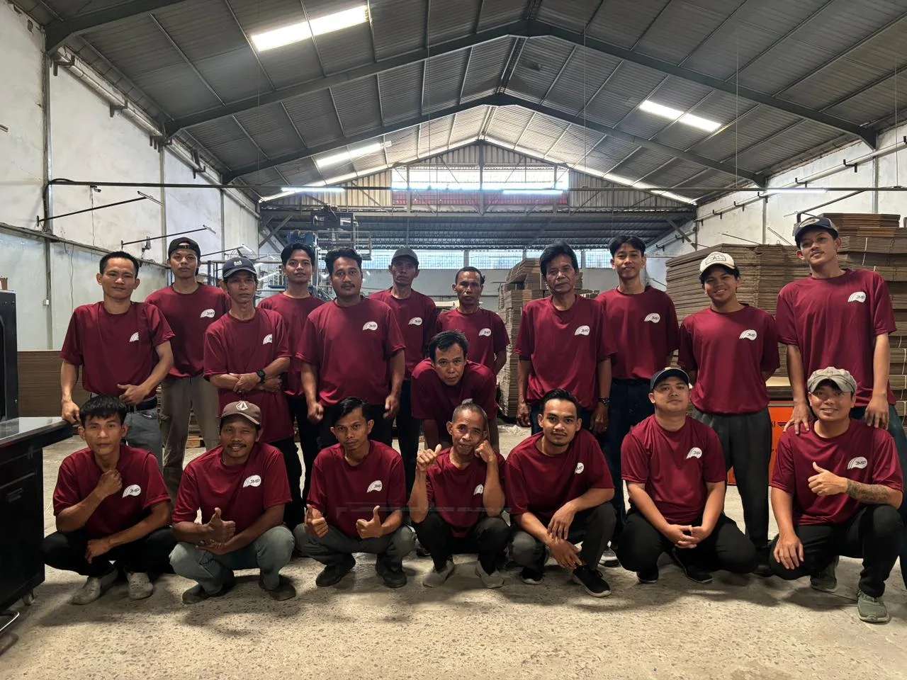 Foto bersama tim divisi carton box berkaus maroon di dalam gudang karton, dengan tumpukan lembaran karton di belakang.