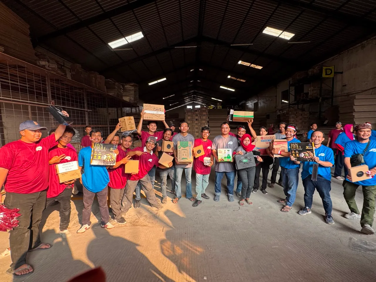 Pembagian doorprize saat hari sudah gelap: dua barisan karyawan berkaus maroon dan biru mengangkat hadiah masing-masing — pan grill, kipas angin berdiri, dispenser air, blender.