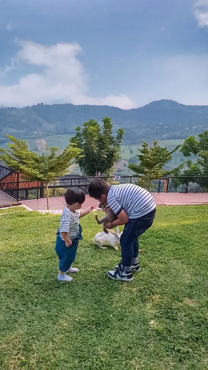 Seorang ayah dan anak memberi makan kelinci dengan latar pegunungan Cianjur.
