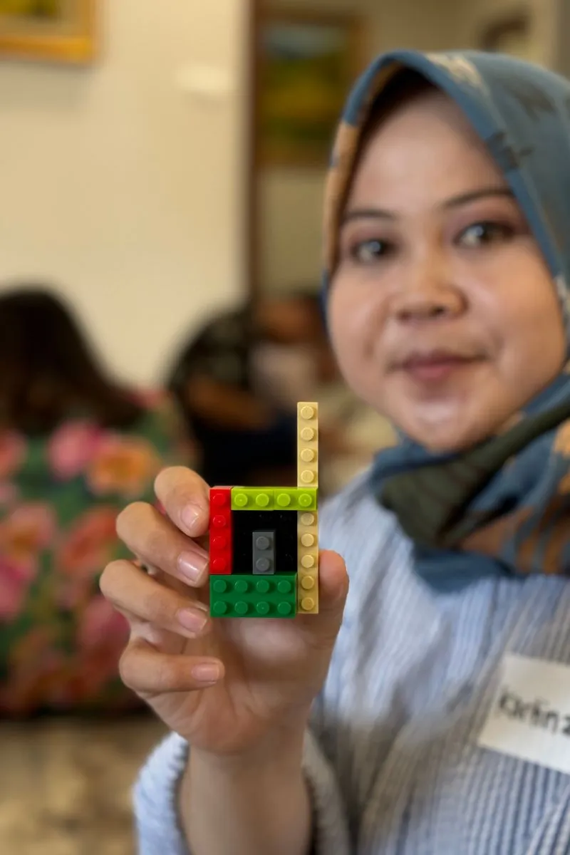 A participant holding up her finished LEGO model to the camera, handwritten name tag on her shirt.